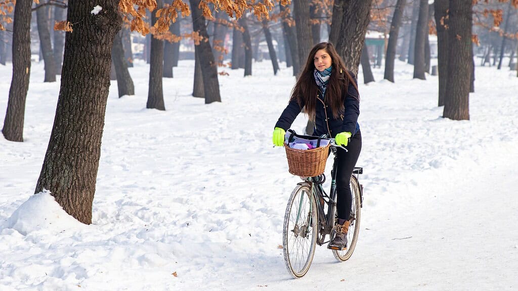 Lány kerékpározik télen, hideg időben, sárga kesztyűben, parkban, hótakaróval borított tájon, környezetbarát közlekedés, téli kerékpározás, téli sport, városi kerékpárút, természet közeli életmód, egészséges életmód, outdoor tevékenység, sport és szabadidő, téli kerékpárút, környezetbarát utazás, sportfelszerelés, túrázás, város és természet kombinációja.